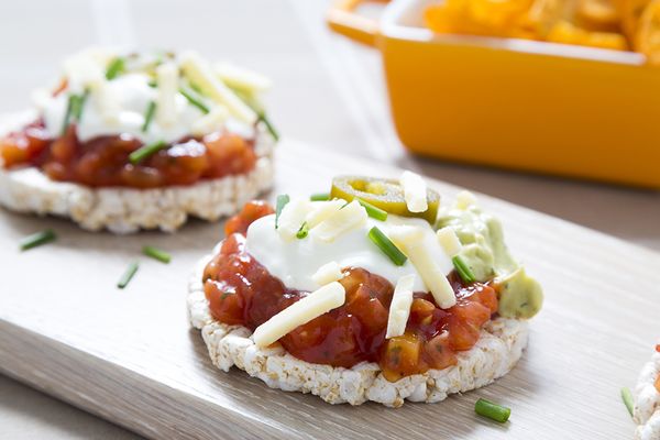 Rice cakes topped with tomato salsa, gaucamole, sour cream, grated cheddar cheese, jalapeno peppers and chopped chives. Tortilla style crisps are in the background