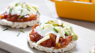 Rice cakes topped with tomato salsa, gaucamole, sour cream, grated cheddar cheese, jalapeno peppers and chopped chives. Tortilla style crisps are in the background